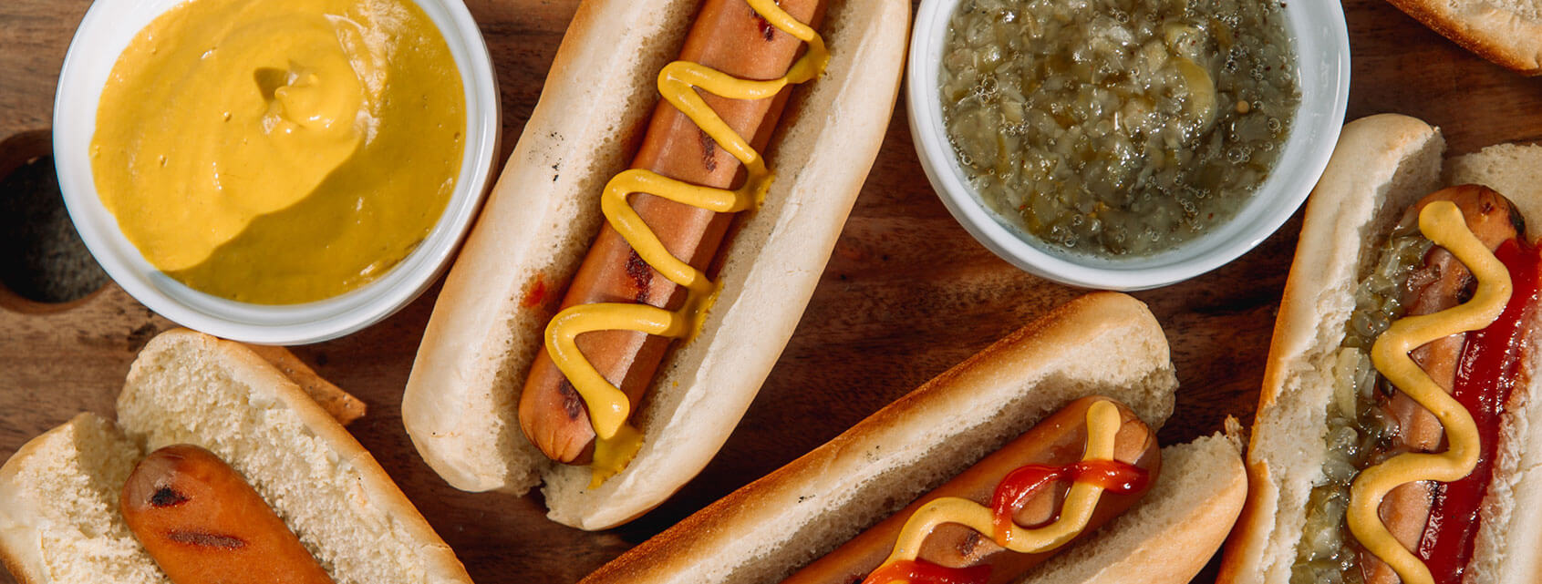 Overheard shot of grilled hot dogs dressed up with ketchup, mustard, and relish next to small bowls of mustard and relish.